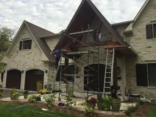 Painter staining the wooden gable of a stone house from a ladder